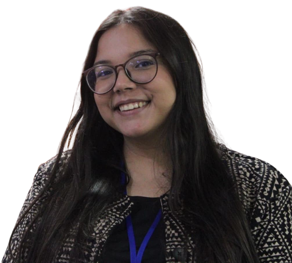 Cynara Maíra. Uma mulher branca de cabelos castanhos escuros usa óculos e está maquiada. Ela sorri para câmera. A mulher usa um blazer estampado preto e bege, com uma regata preta por baixo, com um crachá azul.