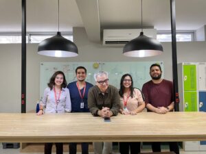 Cinco pessoas possam em frente a uma mesa com dois lustres. No meio está um homem branco de cabelo grisalho que sorri, ele é Jamildo Melo. Todos usam um crachá do Jornal do Commercio em vermelho. São dois homens jovens e duas mulheres jovens brancas. Cynara é uma delas 