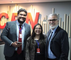 Três pessoas, um homem negro alto de cabelo cacheado preto com um terno cinza e gravata vermelha, ao lado está uma mulher branca baixa de cabelos lisos castanhos, ela usa um blazer estampado marrom e bege, ao lado dela está um homem de cabelo e barba grisalhos com um terno azul marinho. Ao fundo a logo da CBN